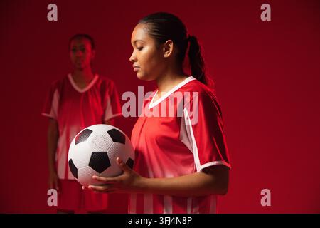 Tossiche africane che tengono un pallone da calcio in studio con illuminazione calda, indossano maglie rosse. Atleta, duo, squadra, attrezzatura, calcio Foto Stock