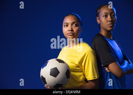 Compagne di squadra afroamericane in piedi schiena contro schiena in studio con maglie e un pallone da calcio. Atletico, lavoro di squadra, ritratto, Foto Stock
