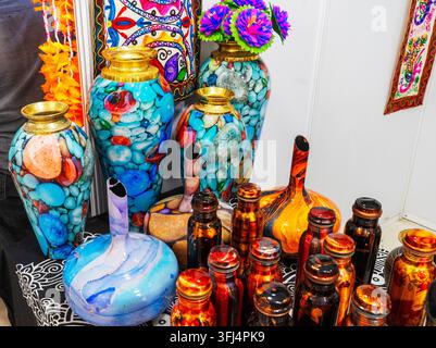 Una colorata esposizione di mercato che mostra oggetti per la casa artigianali e vasi tradizionali. La sezione superiore presenta grandi vasi in ceramica o vetro accattivanti adornati con una stampa a mosaico multicolore che ricorda pietre lisce di fiume e ciottoli oceanici, sormontati da bordi dorati lucidati e fiori finti brillanti. Sotto di essi si trova una selezione di bottiglie d'acqua in rame lucide dipinte artisticamente e flaconi dal collo stretto unici coperti da ricchi motivi a spirale di lacca arancione, rossa e marrone. Foto Stock