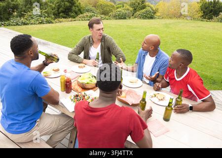 Diversi amici maschi che chiacchierano e afferrano hamburger su un tavolo da picnic in legno sul terrazzo nel giardino. All'aperto, gruppo, sociale, cena, cibo, barbecue, casual Foto Stock
