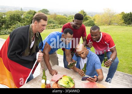 Diversi amici maschi chinati a guardare lo smartphone e tenendo bottiglie di birra verde su un ponte rialzato. Gruppo, all'aperto, picnic, stile di vita, telefono, bandiera, lattina Foto Stock