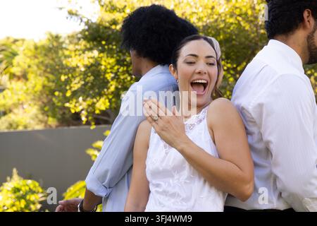 Amici diversi in posa nel cortile, donna che mostra un anello indossando un vestito di pizzo bianco, uomini con camicie. Fidanzamento, giardino, all'aperto, gioielli, celebrazione Foto Stock