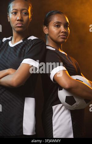 Giocateatrici afroamericane in piedi schiena contro schiena in maglie nere in studio con un pallone da calcio. Duo, atlete, unità, concentrazione Foto Stock