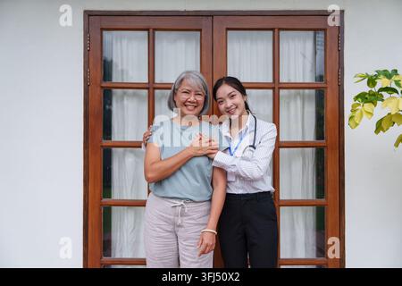 Ritratto di una giovane badante asiatica sorridente che abbraccia una felice donna asiatica anziana davanti a una porta in vetro con cornice di legno a casa. Foto Stock