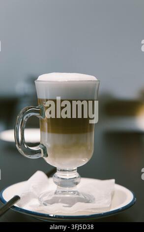 Latte macchiato a strati servito in una tazza di vetro trasparente con latte, caffè e schiuma densa, posizionato su un piattino bianco su uno sfondo sfocato. Foto Stock