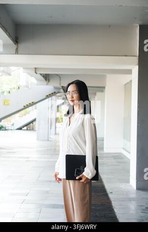 Giovane donna d'affari asiatica con occhiali tiene una cartella nera mentre è in piedi in un corridoio moderno e luminoso dell'ufficio, indossando una camicetta bianca e pantaloni beige Foto Stock