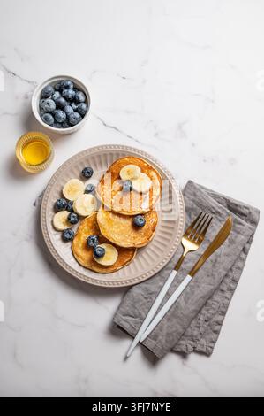 Frittelle con mirtillo e banana su uno sfondo di marmo con posate, miele, tovagliolo e bacche fresche. Un concetto di colazione o brunch vegetariano per Foto Stock