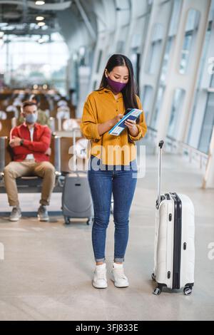 Una donna è in piedi in un terminal aeroportuale tenendo il telefono e i documenti di viaggio mentre un uomo è seduto sullo sfondo, entrambi indossano mascherine Foto Stock