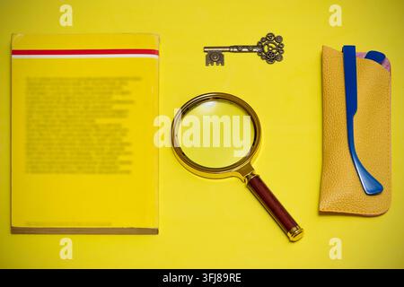 Un libro giallo poggia su una superficie gialla, accanto a una lente d'ingrandimento, una chiave e un paio di occhiali blu con astuccio in pelle gialla Foto Stock