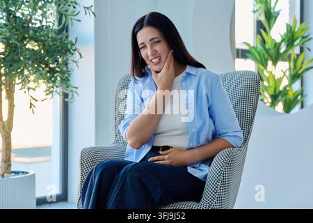 Donna di mezza età che tocca la guancia soffrendo di forti dolori dentali in poltrona Foto Stock
