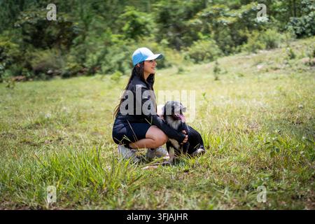 Giovane donna sorridente di profilo, accovacciata e che abbraccia il suo cane in un prato verde Foto Stock