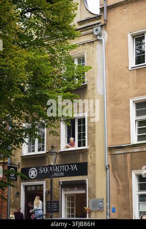 Scatto verticale di una donna con un piccolo cane che guarda fuori da una finestra aperta sopra il ristorante giapponese Sakamoto-ya su una strada storica a Danzica, in Polonia. Foto Stock
