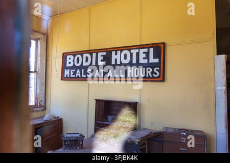 Il cartello dell'hotel Bodie è appeso a un muro giallo sopra mobili vintage, bagagli e un camino all'interno di un edificio storico conservato in California Foto Stock