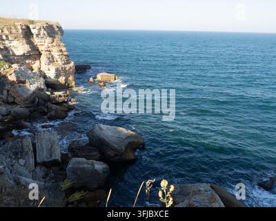 Scogliere di calcare robuste e affioramenti rocciosi lungo una costa drammatica, con mare blu profondo, pilastri marini e onde gentili sotto un cielo sereno Foto Stock