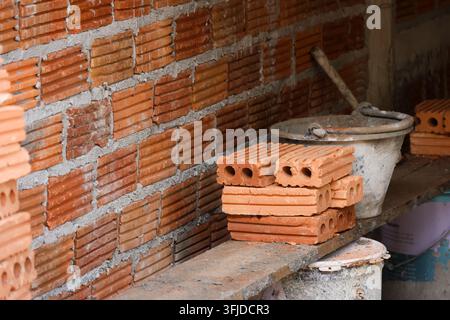 Un muro di mattoni incompiuto e mattoni cavi impilati accanto a un secchio da costruzione in un ambiente da muratura Foto Stock