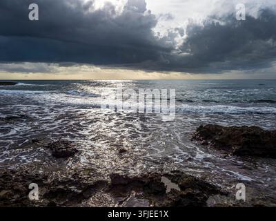 Nuvole scure si librano sopra l'oceano con onde che si infrangono su una costa rocciosa Foto Stock
