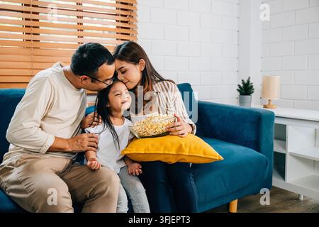Una deliziosa scena familiare si svolge su un divano scanalato mentre si godono il popcorn guardando la TV. padre, madre, figlia e fratello/sorella condividono risate e Foto Stock