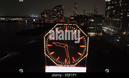 Orologio illuminato sul lungomare di New Jersey City di notte. Skyline con grattacieli sullo sfondo. Bandiera americana sventolante. Ripresa orbitante aerea. Foto Stock