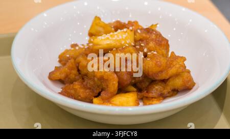 Prima vista di pezzi di pollo croccanti all'ananas ricoperti di una salsa agrodolce e guarniti con semi di sesamo bianchi, serviti in una ciotola bianca. Foto Stock