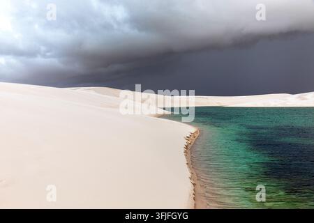 Una vista mozzafiato di una laguna di acqua piovana incontaminata incastonata tra dune di sabbia bianca sotto un cielo tempestoso scuro e drammatico a Lencois Maranhenses Foto Stock