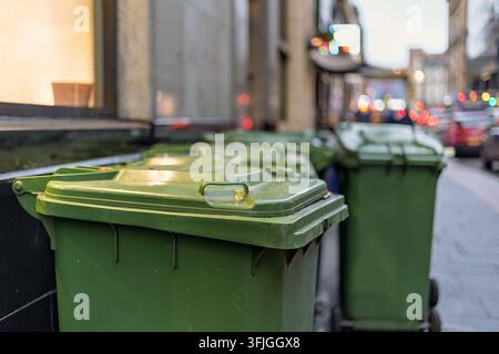 Diverse grandi bidoni della spazzatura in plastica verde con coperchi a battente disposti lungo la facciata di un edificio su una strada urbana trafficata, traffico sfocato e vetrine dei negozi in vista Foto Stock
