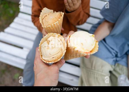 Una vista aerea cattura un adulto che tiene tre coni di gelato su una panchina del parco mentre i bambini giocano nelle vicinanze. Questa gioiosa scena di dolcezza estiva evidenzia la felicità di condividere un dessert dolce all'aperto. Foto Stock
