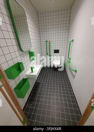 Vista interna di un bagno accessibile con piastrelle bianche sul pavimento e una combinazione di piastrelle bianche e grigio scuro sulle pareti. La stanza include elementi luminosi Foto Stock
