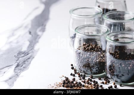 Varietà di pepe nero diverso: pimento, pepe lungo, pepe dei monaci, grani di pepe e polvere macinata in barattoli di vetro su un tavolo da cucina in marmo bianco. Foto Stock