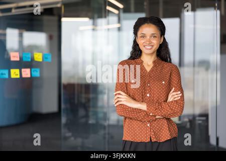 Donna d'affari professionale in piedi in un ufficio moderno con le braccia incrociate, manager donna sicura di sé che dimostra leadership aziendale e successo professionale. Dipendente sorridente in un ambiente di lavoro. Foto Stock