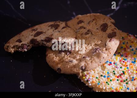 Prima vista di biscotti con gocce di cioccolato e codette colorate disposti su un piano di lavoro scuro, con un biscotto parzialmente morso e briciole per un aspetto invitante Foto Stock