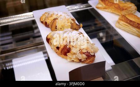 Close up di croissant alle mandorle appena sfornati su piatto rettangolare bianco all'interno di una vetrina da forno in vetro. Pasticceria francese tradizionale ricoperta di aglio affettato Foto Stock