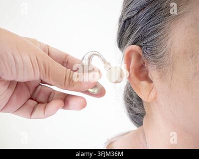 Primo piano di una mano che aiuta un paziente anziano con un apparecchio acustico, sfondo bianco. Foto Stock