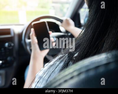 Mockup di una donna che usa il telefono mentre guida al volante. Primo piano all'interno del veicolo di una mano che tiene uno smartphone, volante e interno nero sullo sfondo Foto Stock