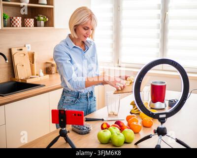 Donna allegra che filma un tutorial sul cibo sano circondata da frutti freschi colorati nella sua cucina. Foto Stock