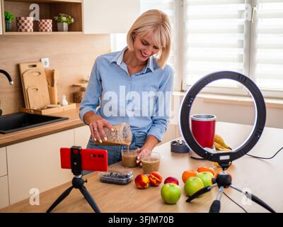 Donna che prepara un frullato fatto in casa circondata da frutti freschi colorati in una cucina moderna e luminosa. Foto Stock