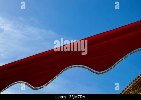 Un'inquadratura a bassa angolazione mostra uno stendardo rosso acceso appeso in alto contro un cielo blu. Piccoli pendenti in metallo simili a monete decorano il bordo dello stendardo. La scena Foto Stock