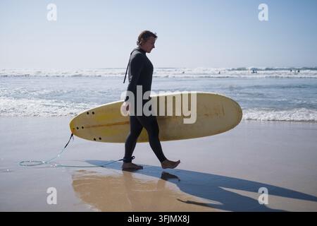 Donna surfista in muta nera che cammina lungo una spiaggia sabbiosa tenendo una tavola da surf Foto Stock