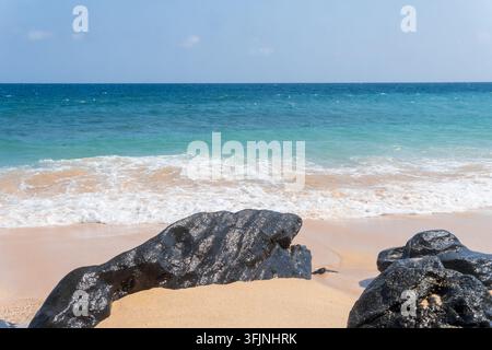 Onde turchesi dell'oceano con schiuma bianca che si infrangono su una riva sabbiosa e rocce vulcaniche nere sotto la luce intensa del giorno su una costa tropicale. Foto Stock