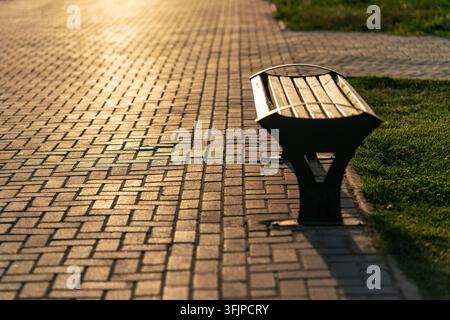 Una panchina di legno solitaria si erge su un sentiero lastricato di pietra in un parco durante un tramonto caldo e dorato. Foto Stock