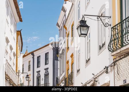 Vista di una strada storica a Évora con edifici bianchi, decorazioni gialle e grigie, balconi in ferro battuto e lanterne appese sotto un cielo blu terso a Évora Foto Stock
