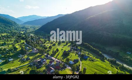 Un insediamento soleggiato si estende lungo una valle verde ai piedi di un vasto pendio. Strade, campi e cottage costeggiano il fiume mentre montagne boscose circondano l'orizzonte Foto Stock