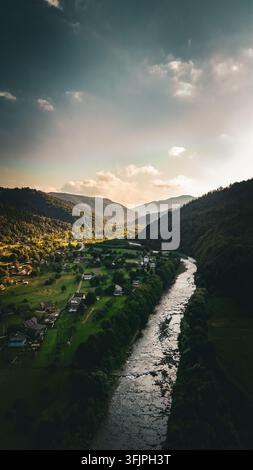 Una scena fluviale verticale attira lo sguardo a monte tra montagne in ombra. I riflessi luccicano sulla corrente mentre piccole case e prati costeggiano la riva Foto Stock