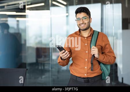 Giovane uomo d'affari che utilizza uno smartphone in un ufficio moderno, uomo professionale con uno zaino in piedi a una scrivania per attività di comunicazione aziendale e connettività digitale durante una giornata lavorativa impegnata. Foto Stock