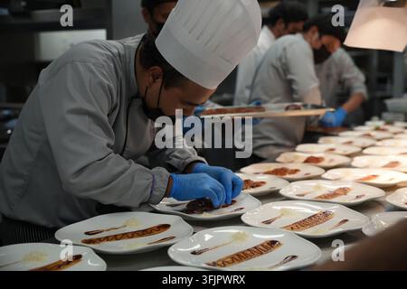 Una squadra di chef professionisti in uniforme che lavorano efficientemente in fila per servire più piatti durante un servizio di banchetto affollato. Foto Stock