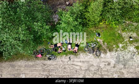 Vista a droni di ciclisti di montagna che si riposano ai margini di una foresta accanto a un sentiero di ghiaia. Le biciclette sono sparpagliate mentre i compagni di squadra si rilassano sull'erba godendosi l'ombra Foto Stock