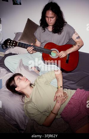 Uomo che suona la chitarra acustica per una donna rilassata a letto a casa Foto Stock