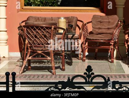 Sedie di vimini con cuscini marroni circondano un piccolo tavolo di vetro accanto a un muro di terracotta, una fioriera frondosa e una ringhiera decorativa che proiettano ombre Foto Stock
