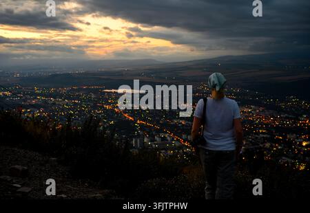 Una vista posteriore di una fotografa in piedi su un punto elevato, che guarda una città estesa al crepuscolo. Indossa una maglietta bianca, grigia Foto Stock