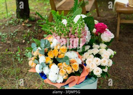 Un lussureggiante mazzo misto disposto in un contenitore decorativo e mostrato all'aperto tra gli alberi. La composizione presenta fiori rosa, bianchi, gialli, arancioni e Foto Stock