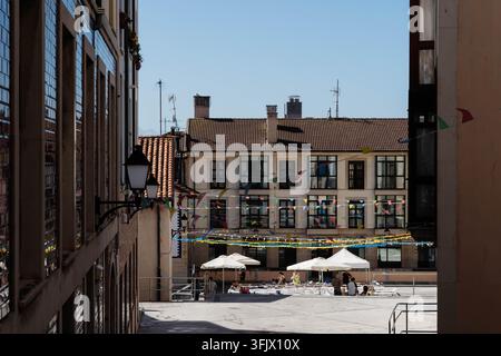 Gijon, Spagna – 19 settembre 2026: striscioni festivi attraversano una piazza di pietra illuminata dal sole dove le persone si radunano sotto ombrelloni bianchi su una terrazza di un caffè. Fiesta Foto Stock